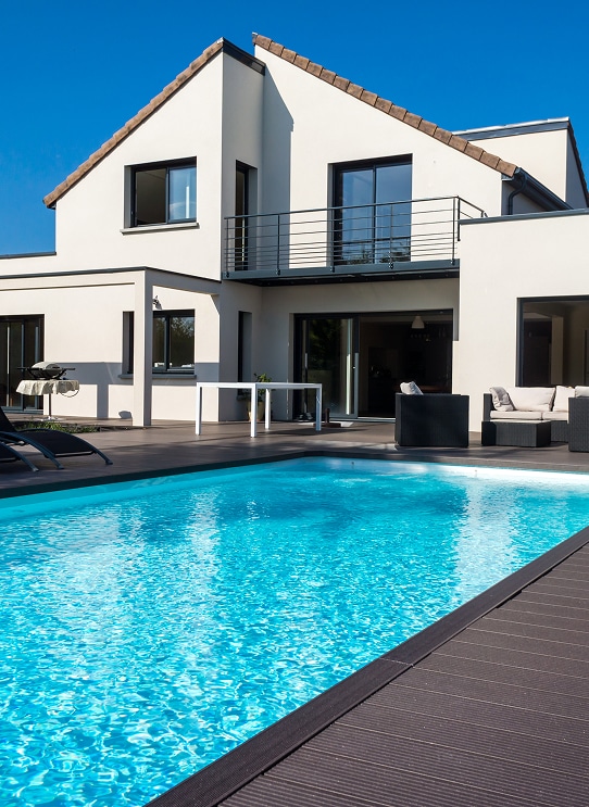 Maison moderne blanche avec toit tuile, piscine turquoise et grande terrasse en bois sombre. Mobilier extérieur.