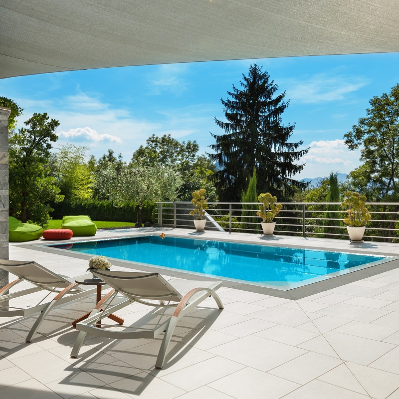 Piscine extérieure moderne avec terrasse carrelée, transats, vue sur jardin luxuriant et arbres sous voile d'ombrage.