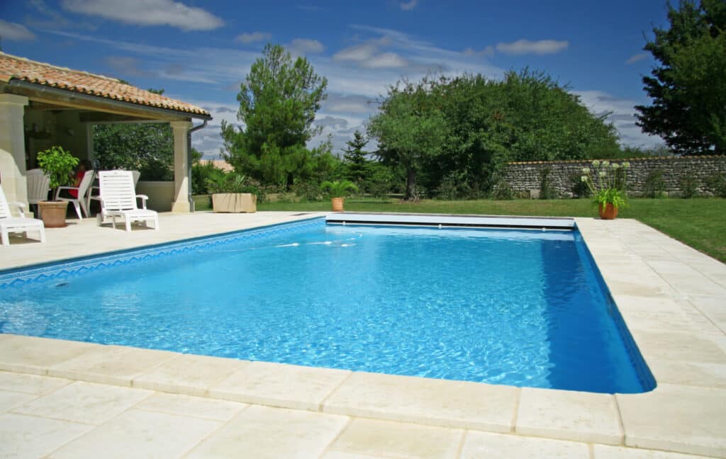Piscine bleue ensoleillée avec terrasse en pierre claire, chaises longues blanches et jardin verdoyant. Un cadre extérieur paisible.