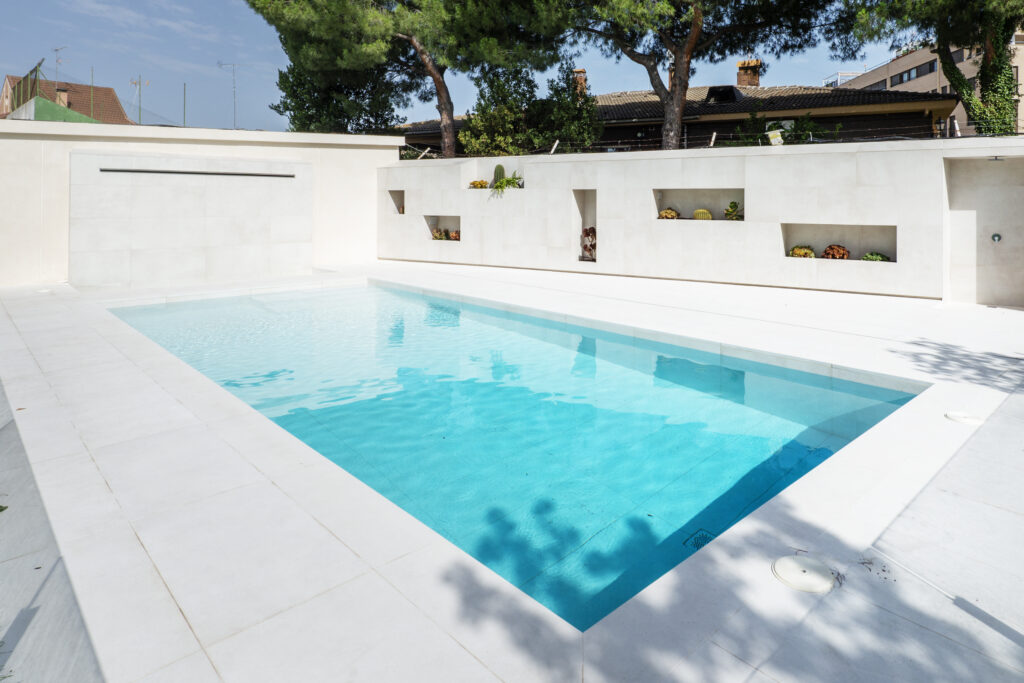 Grande piscine rectangulaire moderne à l'eau bleue claire, entourée de carrelage blanc, avec un mur élégant, cascade et niches végétales.