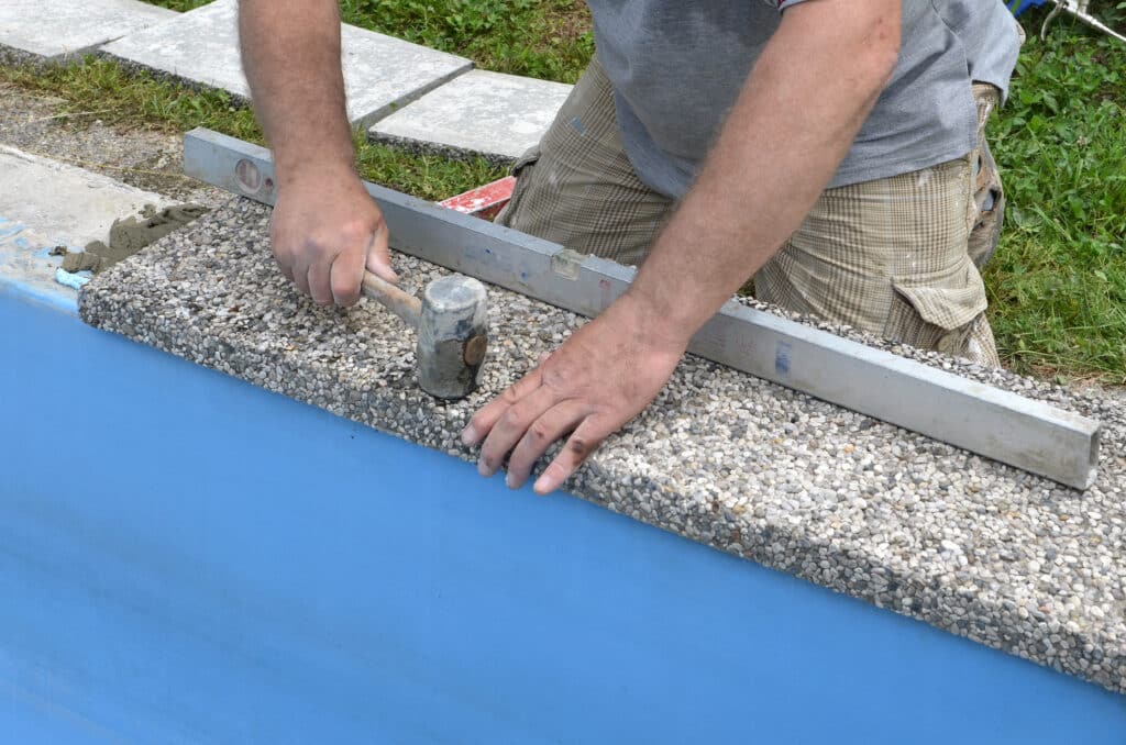 Ouvrier posant une bordure en béton de galets avec un marteau et un niveau, le long d'un bassin bleu de piscine.